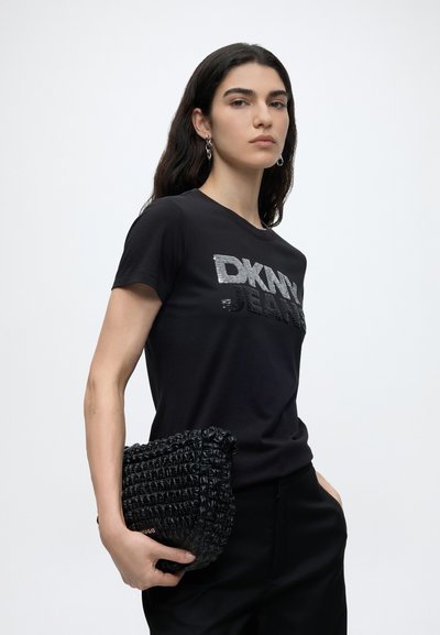 Млада жена с дълга тъмна коса, носеща черна тениска DKNY Jeans и държаща текстурирана черна клъч чанта на едноцветен фон.