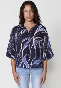 Camicia blu navy con vestibilità ampia, maniche corte e chiusura a bottoni. Presenta un motivo a foglia in diverse tonalità di blu e bianco sporco. Tessuto leggero.