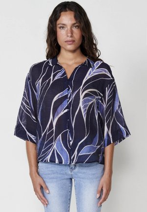 Camicia blu navy con vestibilità ampia, maniche corte e chiusura a bottoni. Presenta un motivo a foglia in diverse tonalità di blu e bianco sporco. Tessuto leggero.