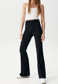 Młoda kobieta ubrana w biały crop top, czarne wysokie szerokie spodnie, białe trampki oraz kolorowe pierścionki na obu rękach.