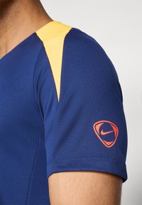 Modra športna majica z rumenimi delom na ramenih. Teksturiran material z majhnim oranžnim Nike logotipom na rokavu.