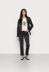 Jaqueta de couro preta sobre uma t-shirt gráfica rosa com texto verde e um morango. Combinada com jeans pretos e ténis preto/brancos.