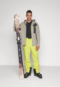 Skio equipment met een mannelijke model die een paar ski's vasthoudt. Hij draagt een lichtgrijze jas, een zwart shirt en felgele broek. De skischoenen zijn zwart.