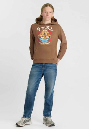 Young person wearing brown hoodie with ramen bowl and dog graphic, blue jeans, and white sneakers standing against plain background.