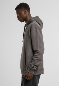 Grå hettegenser laget av mykt stoff, med en frontlomme, snøring på hetten og ribbestrikkede mansjetter. Enkel design med minimal detaljering.