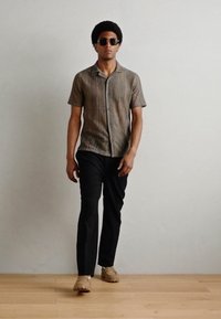 Cămașă cu mânecă scurtă, cu un model în nuanțe de marron, asortată cu pantaloni negri și încălțăminte bej. Modelul poartă ochelari de soare și stă pe un podea din lemn.