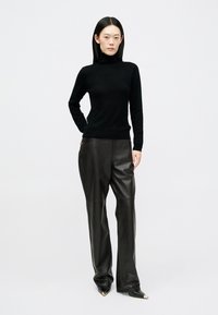 Mulher com cabelo preto liso, vestindo um suéter de gola alta preto, calças de couro pretas largas e sapatos pretos de bico fino com pontas douradas, em pé.