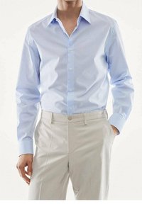 Homme portant une chemise habillée à manches longues à carreaux bleu clair rentrée dans un pantalon beige, une main dans la poche, sur un fond uni.