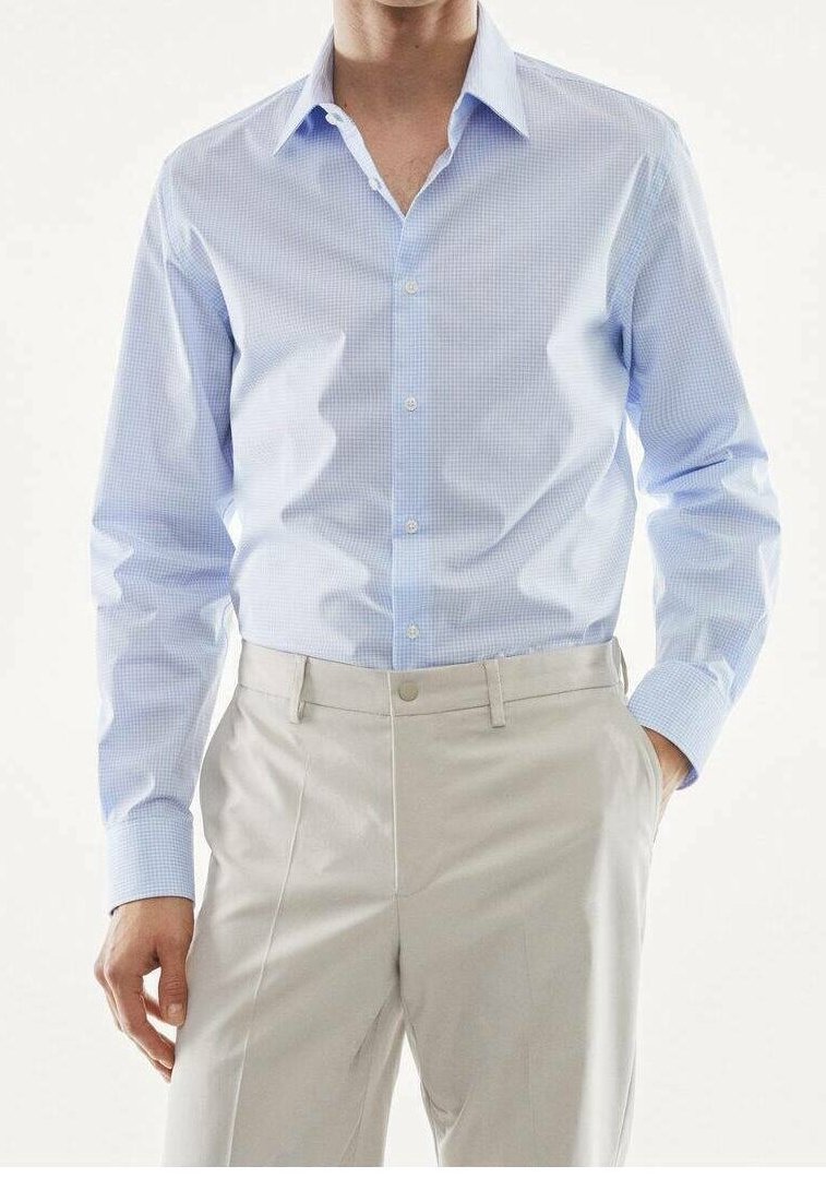 Homme portant une chemise habillée à manches longues à carreaux bleu clair rentrée dans un pantalon beige, une main dans la poche, sur un fond uni.
