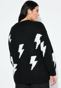 Frau in schwarzem Strickpullover mit weißen Blitzmustern, seitlich vor einem schlichten weißen Hintergrund stehend.