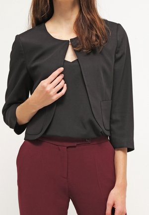 Nainen pukeutunut mustaan lyhytvartiseen takkiin mustan paidan päällä, yhdistettynä korkeavyötäröisiin viininpunaisiin housuihin yksiväristä taustaa vasten.