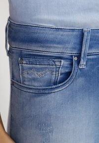 Los jeans de mezclilla azul claro presentan un bolsillo frontal con una costura curva y un patrón de puntadas decorativas, mostrado en una vista en primer plano.