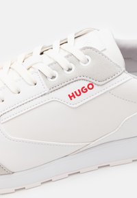 Hvite sneakers med glatt skinn og semsket lær detaljer, rund tå, seks snøreøyler, og rødt "HUGO"-logo på siden.