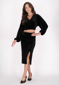 Schwarzes Samt-Cropped-Top mit langen Ärmeln und V-Ausschnitt, kombiniert mit einem schwarzen Samt-Midirock mit seitlichem Schlitz und Knopfdetails.