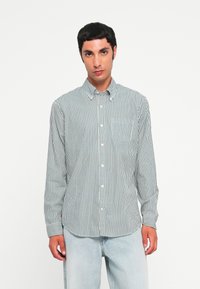 Chemise en coton à manches longues avec un motif rayé vertical vert et blanc, col boutonné et poche avant. Associée à un pantalon en denim bleu clair.