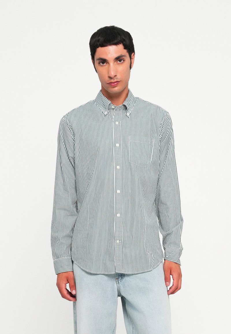 Chemise en coton à manches longues avec un motif rayé vertical vert et blanc, col boutonné et poche avant. Associée à un pantalon en denim bleu clair.