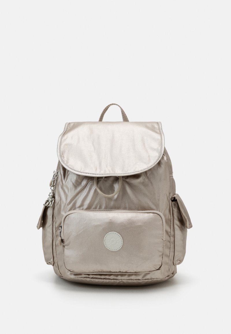 Metaliczny beżowy plecak z klapą, górnym uchwytem, przednią kieszenią na zamek błyskawiczny i kieszeniami bocznymi. Posiada okrągłe logo z przodu.