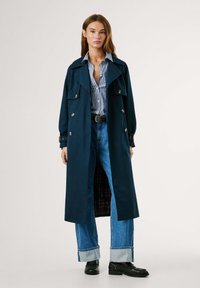 Manteau long navy à coupe décontractée, avec de grandes poches avant, des boutons dorés et des poignets retroussés. Chemise rayée bleue et blanche en dessous.