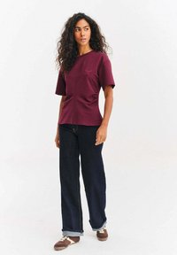 Burgundské tričko s krátkými rukávy a bočními volánky, kulatý výstřih; v kombinaci s tmavými džínami širokých nohavic a hnědými teniskami s bílými podrážkami.