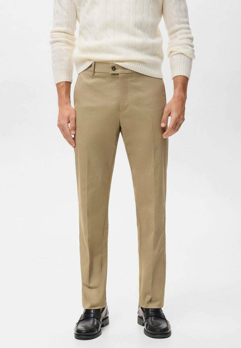 Beige op maat gemaakte broeken met een vlakke voorkant, rechte pijpen en subtiele plooidetails. Gecombineerd met zwarte instappers en een crèmekleurige trui.