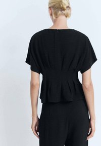 Una donna con i capelli biondi, che indossa un top di stile peplum nero, si volta mostrando il suo tessuto testurizzato e la silhouette rilassata su uno sfondo chiaro.
