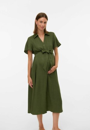 Zwangere vrouw in een knielang olijfgroen jurkje met korte mouwen en een gestrikte taille, die zachtjes haar buik vasthoudt, staande op een witte achtergrond.
