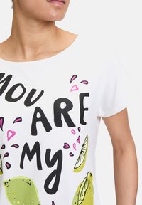 Biała bawełniana koszulka z odważnym czarnym napisem "JESTEŚ MOJĄ" w zabawnym wzorze z zielonymi cytrynami i różowymi sercami. Krótkie rękawy i okrągły dekolt.