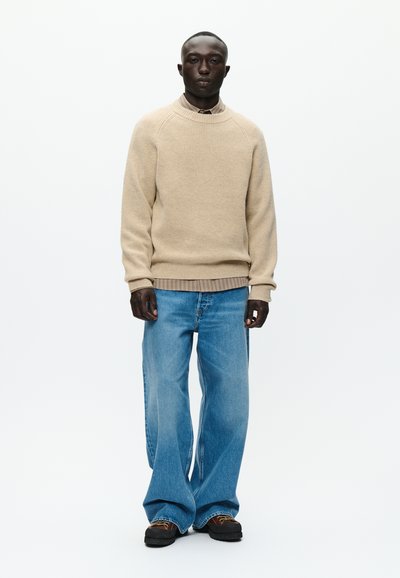 Bežasti rebrasti pulover s okruglim izrezom, uparen s širokim svijetloplavim trapericama. Model nosi tamnu obuću i stoji pred bijelom pozadinom.