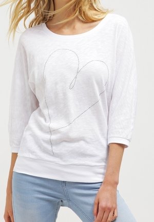 Vrouw met een wit shirt met lange mouwen waarop een gestikte omtrek van een want is afgebeeld, en lichtblauwe spijkerbroek, gezoomd op halverwege de dijen.