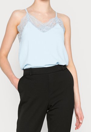 Frau trägt hellblaues Spitzen-Top, in hochtaillierte schwarze Hose gesteckt, Hand in der Tasche, vor schlichten Hintergrund.