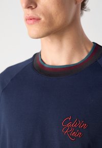 Tamsiai mėlynas medvilninis džemperis su įmegztu apvaliomis iškirptėmis, puoštais raudonais, žydrais ir juodais dryžiais, bei raudonu siuvinėtu "Calvin Klein" logotipu.