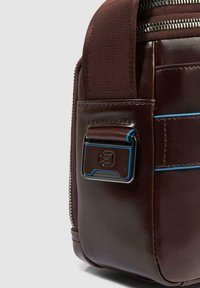 Angolo di borsa in pelle marrone scuro con cerniera, tracolla spessa, bordatura blu e chiusura metallica con logo impresso.