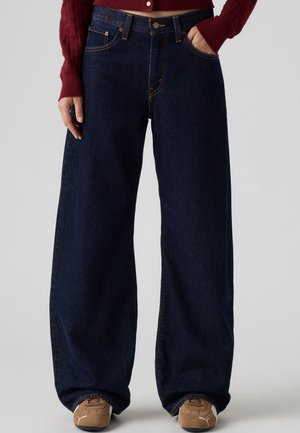 Personne portant un jean large bleu foncé, des baskets marron et un pull rouge court avec une main dans la poche.