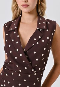 Top senza maniche marrone con scollatura a V profonda, caratterizzato da grandi pois crema e colletto a punta. Tessuto liscio con taglio sartoriale.