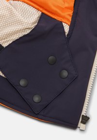 Detailní záběr na roh bundy, který ukazuje tmavě modrou látku se čtyřmi černými patentkami, bílým zipem, oranžovou podšívkou a béžovou vzorovanou vnitřní látkou.