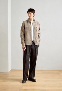 Chaqueta con cuello en un patrón de cuadros beige y marrón, con dos bolsillos en el pecho, combinada con pantalones oscuros y zapatos negros sobre un suelo de madera.