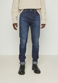 Vaqueros de mezclilla azul oscuro con un corte slim, con cierre de botones, cinco bolsillos y puños remangados. Combinados con botas negras hasta el tobillo.