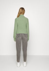 Mujer con cabello largo y rubio, vistiendo un suéter de cuello alto verde, jeans grises y zapatillas blancas, de pie frente a una pared blanca.