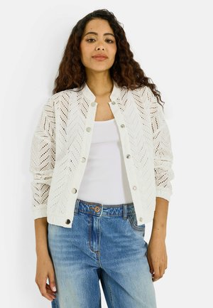 Mujer con cabello largo y rizado que lleva una chaqueta blanca con botones y estampado sobre una camiseta blanca sin mangas y vaqueros azules.
