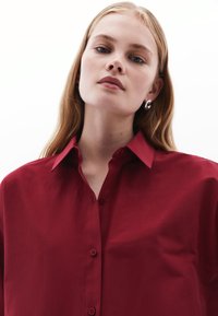 Rotes Hemd mit Knopfleiste und spitzem Kragen, aus glattem Stoff und in lockerer Passform. Dezente Knöpfe und klare Designdetails sind sichtbar.