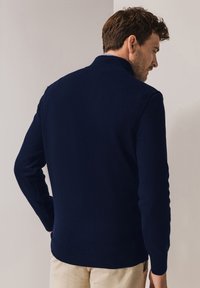 Marineblauwe gebreide trui met een hoge kraag, lange mouwen en geribbelde manchetten, ontworpen voor een nauwsluitende pasvorm, gecombineerd met lichtgekleurde broek.