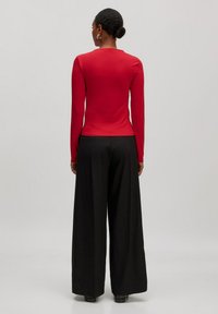 Rotes Langarmoberteil kombiniert mit schwarzen, weit geschnittenen Hosen, die eine figurbetonte Silhouette und eine glatte Textur aufweisen, von hinten betrachtet.