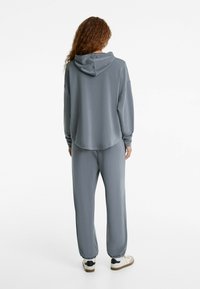 Hanorac gri cu glugă ajustabilă, croială supradimensionată și manșete ribbate, asortat cu pantaloni jogging gri, cu glezne elastice și o textură netedă.