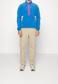 Pullover in pile blu con patta dei bottoni rosa e tasche laterali arancioni, abbinato a pantaloni beige e sneakers grigie. Texture liscia.
