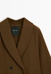 Brązowy płaszcz z mieszanki wełny z kołnierzem z wyłogami, podwójnym zapięciem oraz subtelną teksturą. Wewnątrz znajduje się czarna metka.