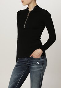 Czarny, prążkowany top z długim rękawem z zamkiem błyskawicznym z przodu, z metalowymi elementami, dopasowany fason i miękka tekstura. W połączeniu z przetartymi niebieskimi dżinsami.