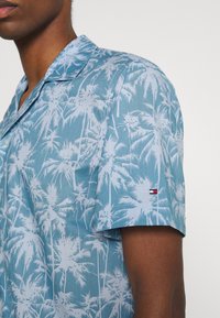Camisa de manga corta en tela azul claro con patrones de palmeras blancas, con cuello en pico y un pequeño logo en la manga.