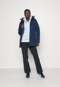 Navy blue waterproof jacket with a zip front, large hood, and multiple pockets, layered over a light blue hoodie and black pants.