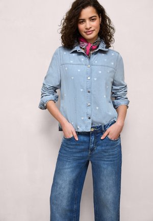 Frau trägt ein hellblaues Jeanshemd mit Herzmuster, hochgekrempelte Ärmel, blaue Jeans und ein rosa gemustertes Halstuch, Hände in den Taschen.