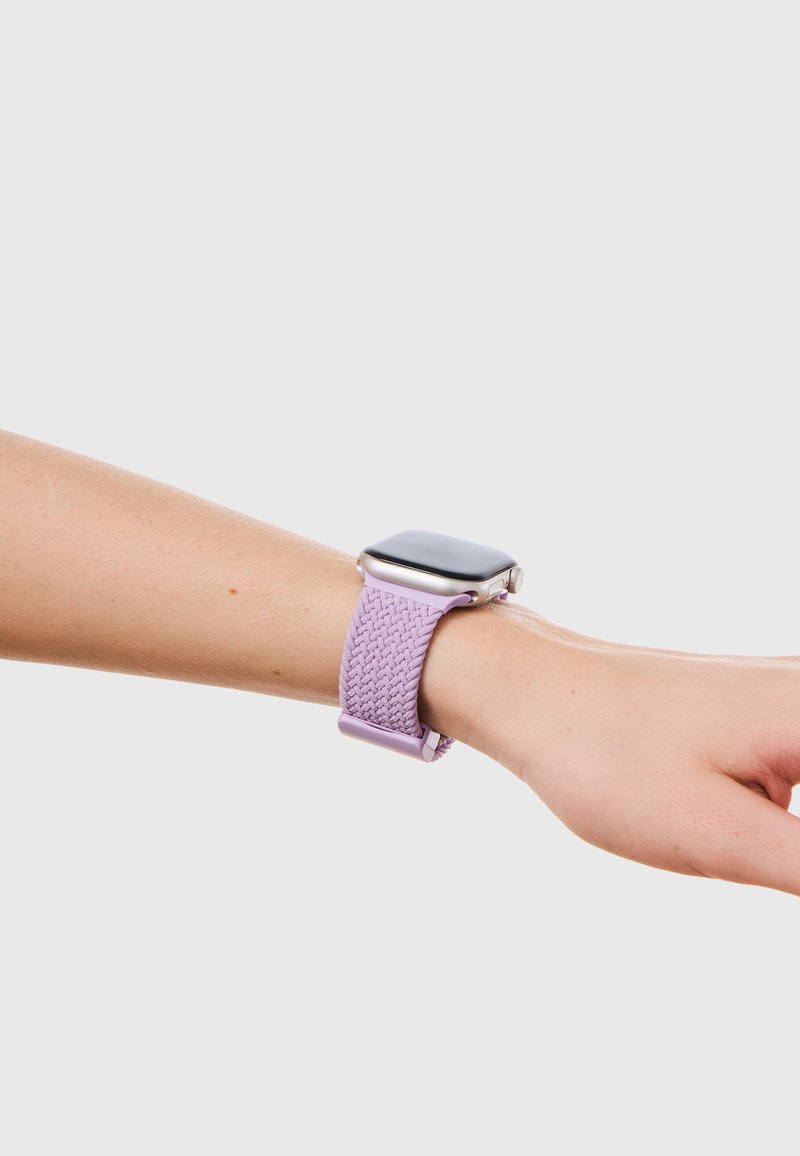 Καρπός με ένα smartwatch με ένα ανοιχτό μοβ πλεκτό λουράκι σε ανοιχτό τόνο δέρματος σε απλό γκρι φόντο.